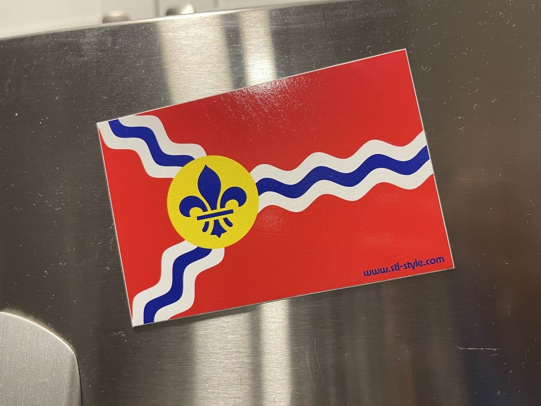 A refrigerator magnet of the City of St Louis flag with a wavy blue and white stripe and a yellow circle featuring a blue fleur-de-lis on a red background, all placed on a stainless steel refrigerator door. 