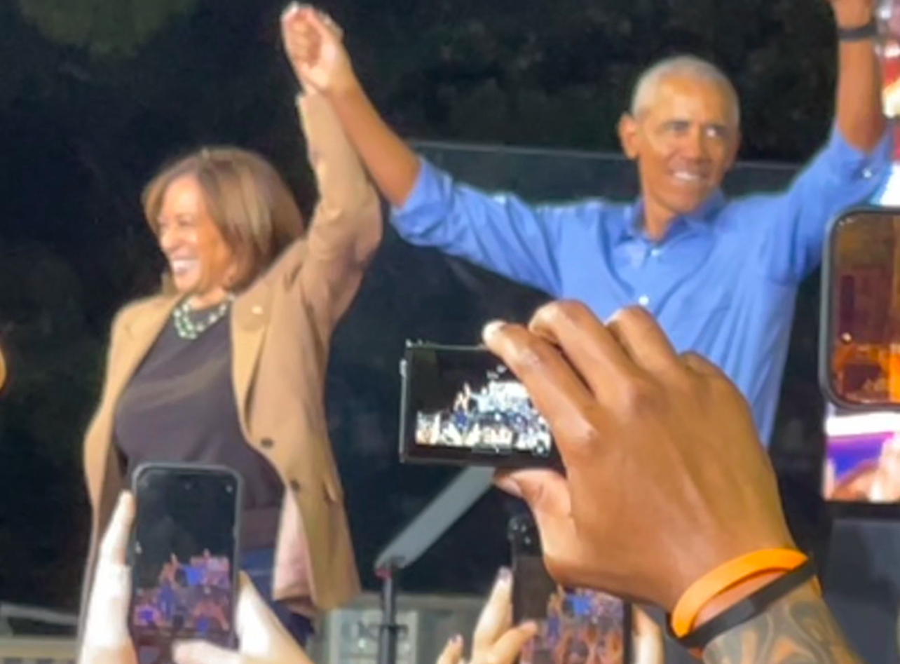 Two people (Harris and Obama) are standing on stage holding hands, surrounded by a crowd taking photos.