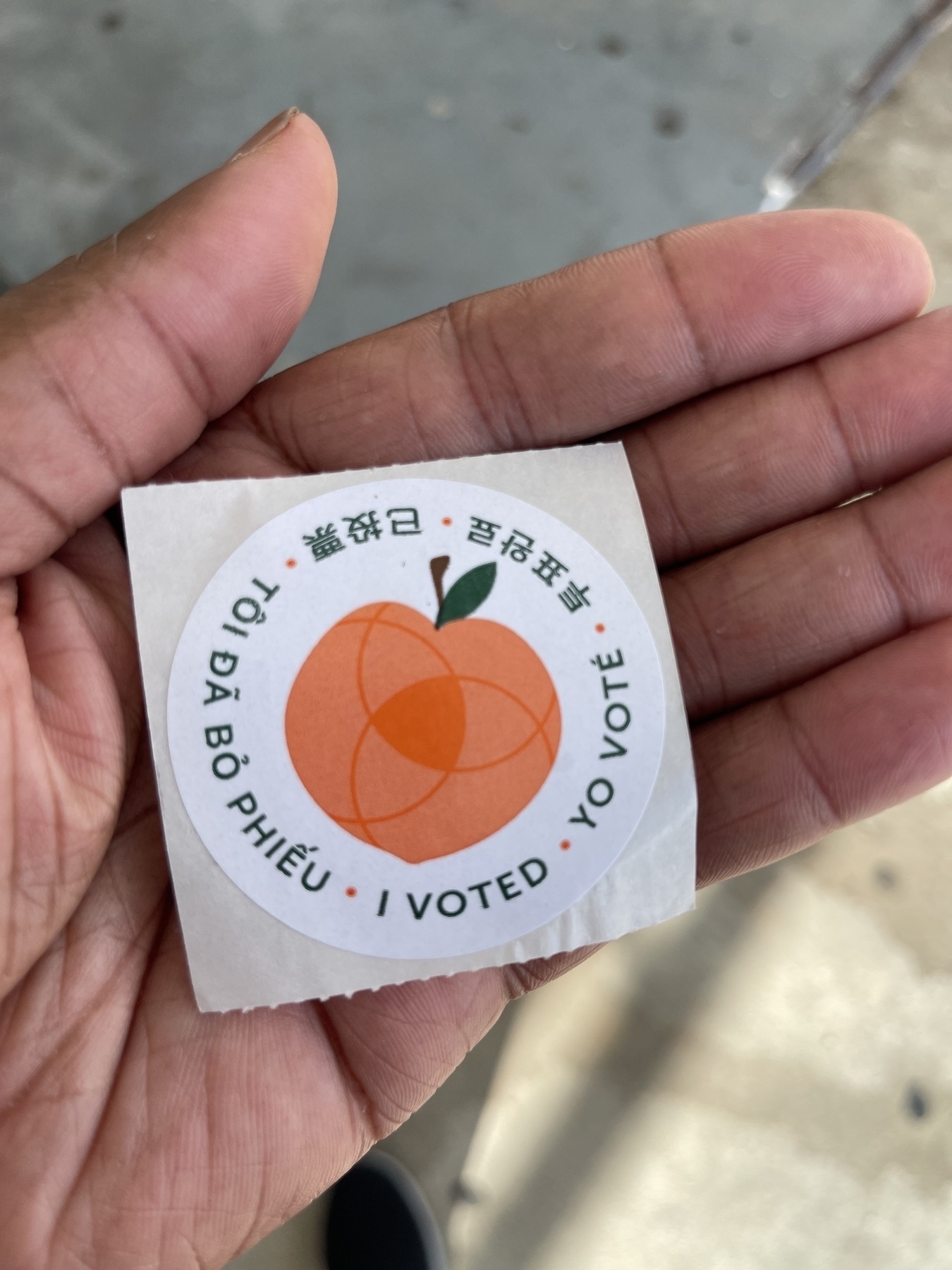 A hand is holding an "I Voted" sticker featuring a peach illustration and multiple languages.